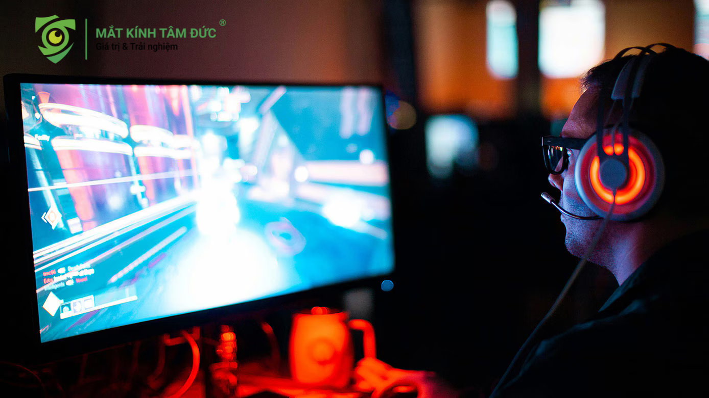 Cận cảnh nam game thủ đeo kính với tai nghe phát sáng đang chơi game trong môi trường thiếu sáng, ánh sáng màn hình rất chói đừng để mắt bạn bị ảnh hưởng hãy làm giảm mỏi mắt game thủ khi chơi